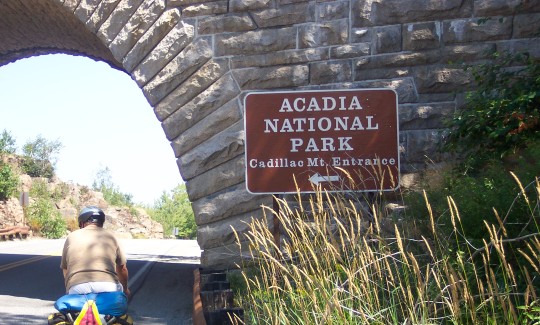 Don riding under a bridge with sign, 'Acadia NP, Cadillac Mt. Entrance'