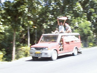 Small truck with passengers on roof.  One holds his coat in the air as a greeting.